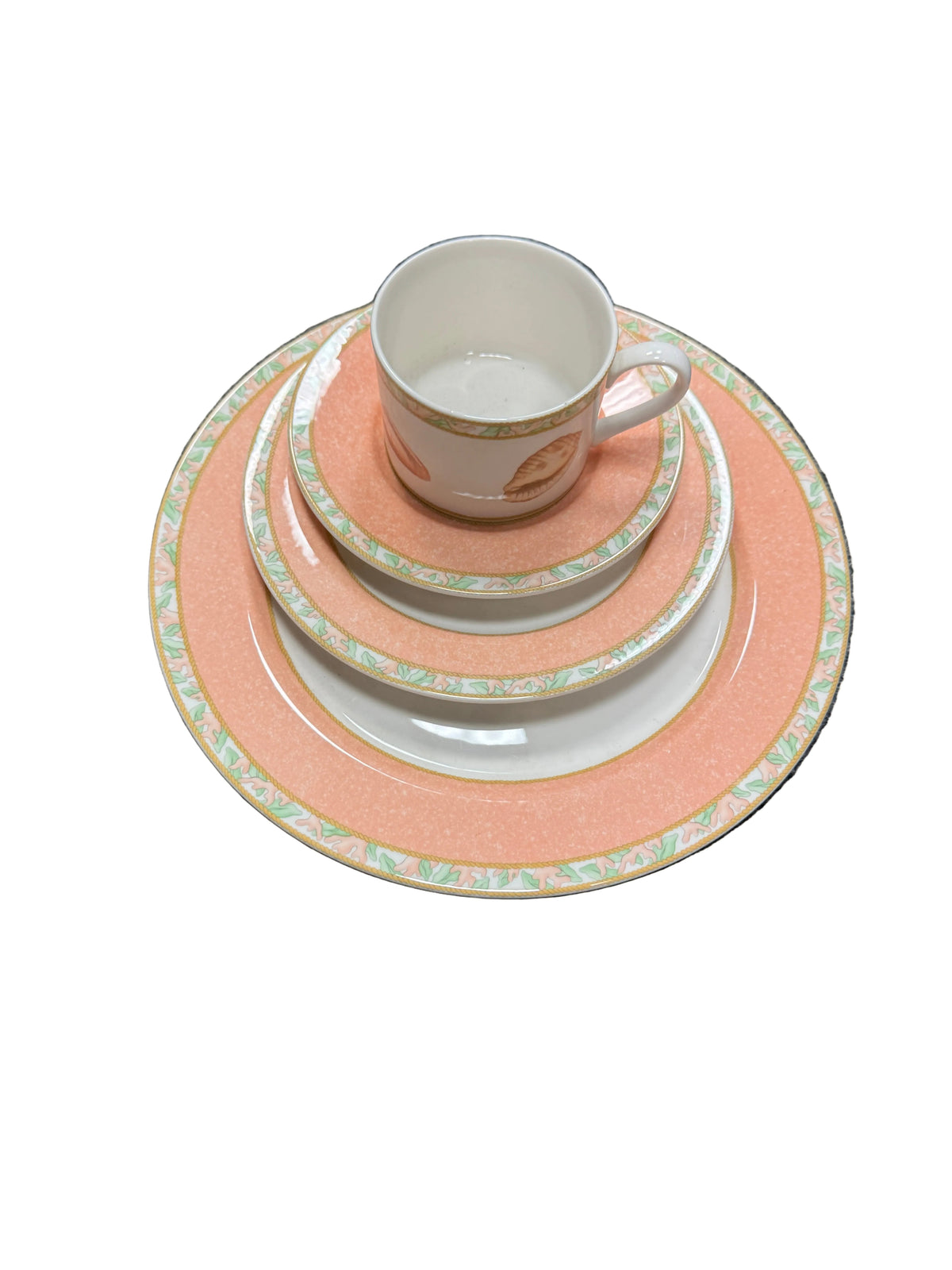 A stack of ceramic tableware including a teacup, saucer, plates, and charger plates in a peach and white color scheme with a green and gold patterned rim, set on a gray carpeted floor.