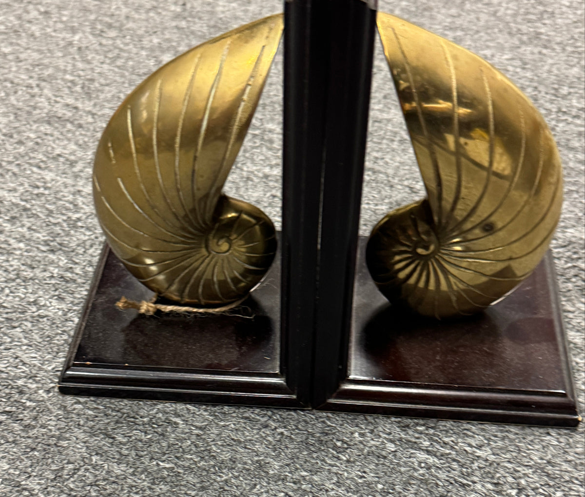 A pair of decorative bookends shaped like polished gold nautilus shells on dark wooden bases, photographed on a gray carpeted floor.
