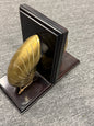A decorative pair of bookends featuring polished gold-toned leaf-shaped tops and dark brown wooden bases, shown standing on a gray carpeted floor.