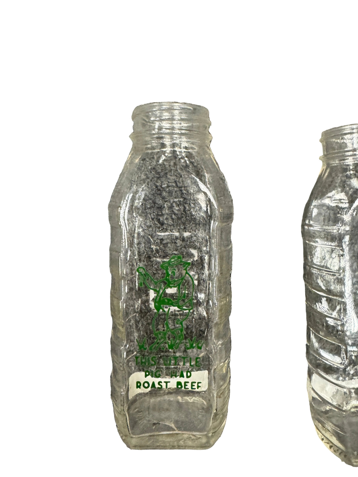 A clear glass bottle with embossed green text reading 'This Little Pig Had Roast Beef' lying on a gray textured carpet, with another similar bottle partially visible on the right.