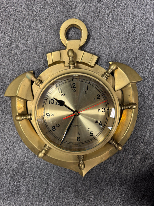 A brass nautical watch or clock with a circular dial, black numerals, and decorative screw-like accents, photographed from above on a gray textured surface.