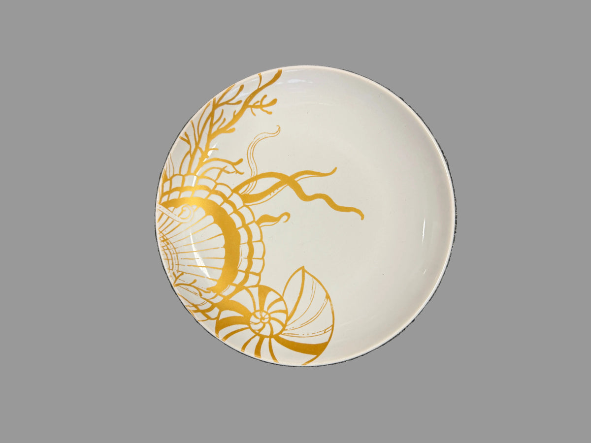 A round white ceramic plate with a gold seashell and coral-like line art design, photographed from above on a textured gray surface.