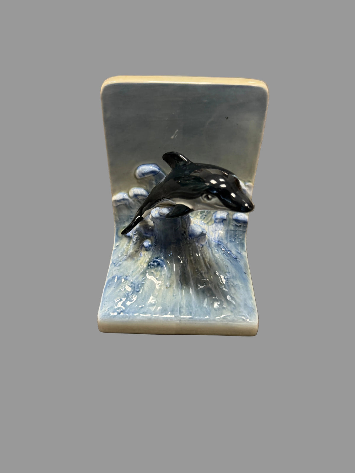 A ceramic bookend featuring a black and white painted dolphin jumping over blue and white waves, set against a gray carpeted floor.
