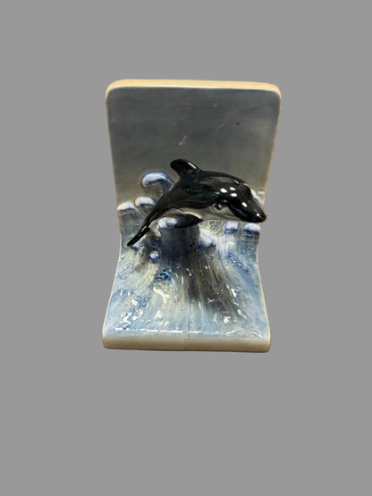 A ceramic bookend featuring a black and white painted dolphin jumping over blue and white waves, set against a gray carpeted floor.